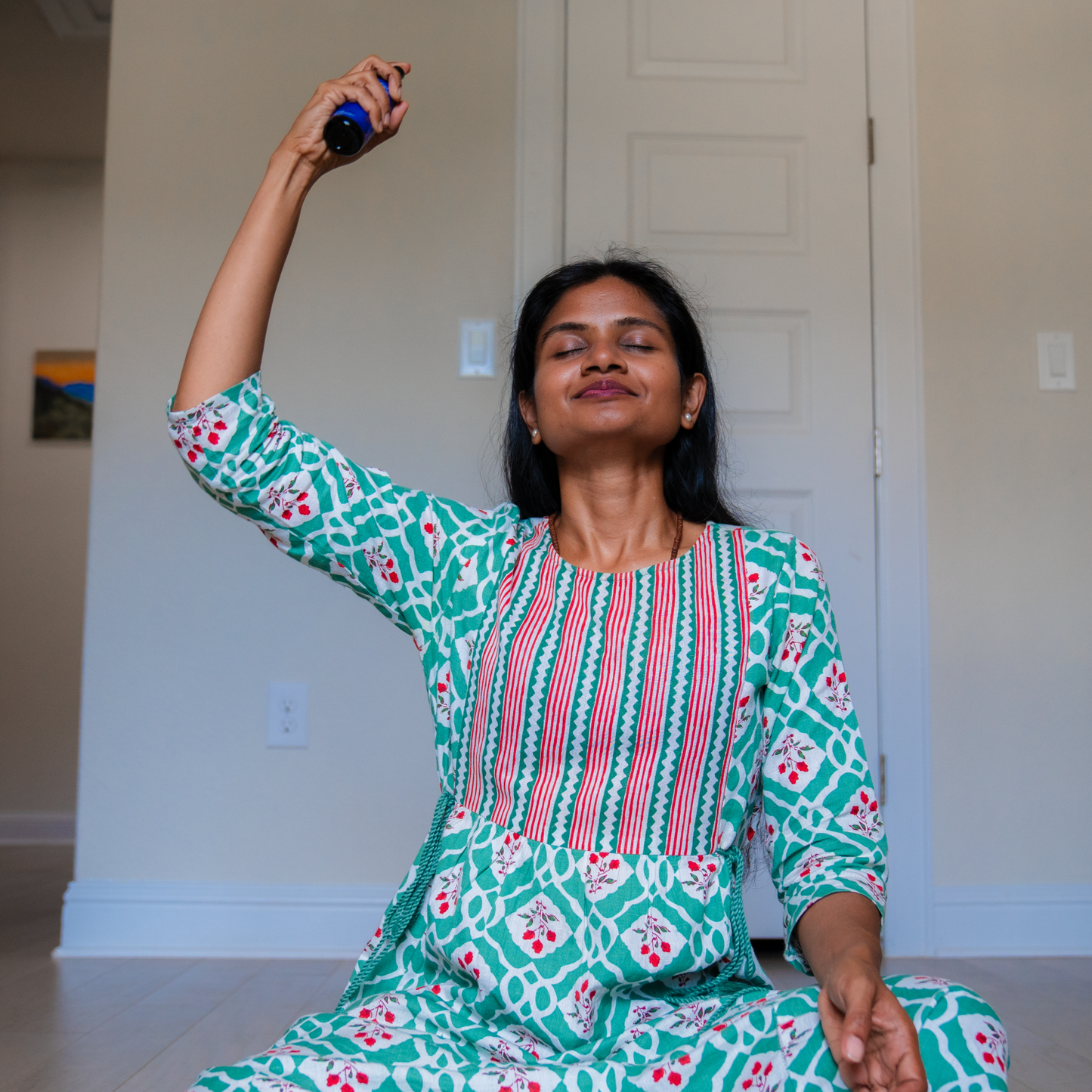 Woman sitting cross-legged, eyes closed, spraying LOTUSWEI Luscious Embodiment Aura Mist above her head indoors