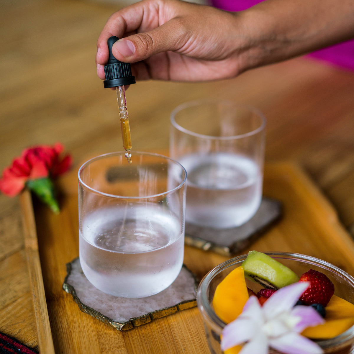 Hand adding dropper drops to a glass of water, a red hibiscus bloom and a fruit bowl with an orchid garnish nearby.
