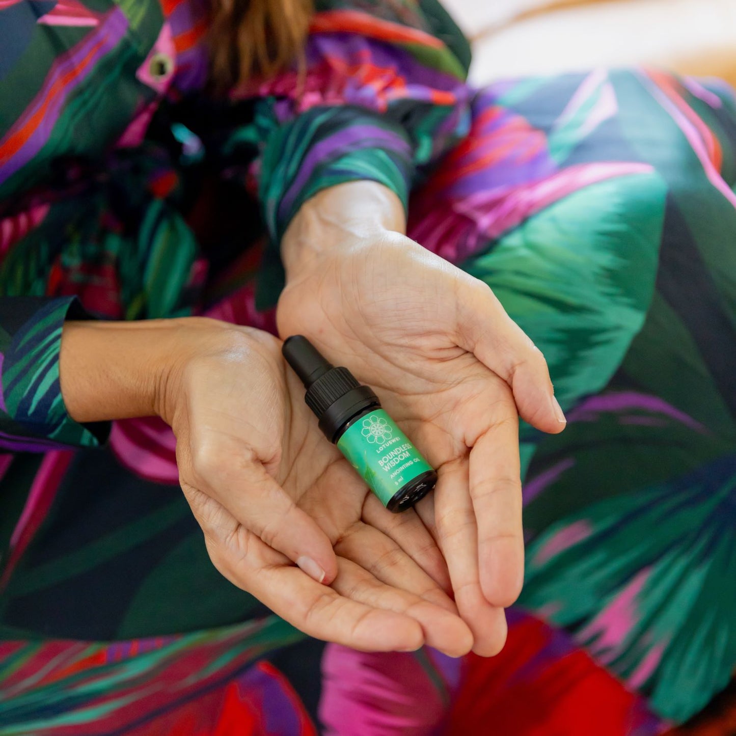 Hands cupping a small LOTUSWEI Boundless Wisdom Anointing Oil bottle against a colorful print sleeve