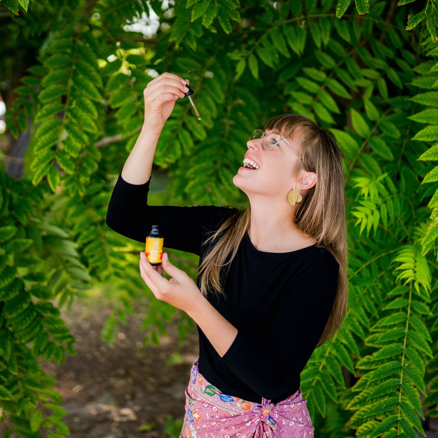 Woman laughing while lifting an eyedropper above a LOTUSWEI Inspired Action Elixir bottle in green foliage