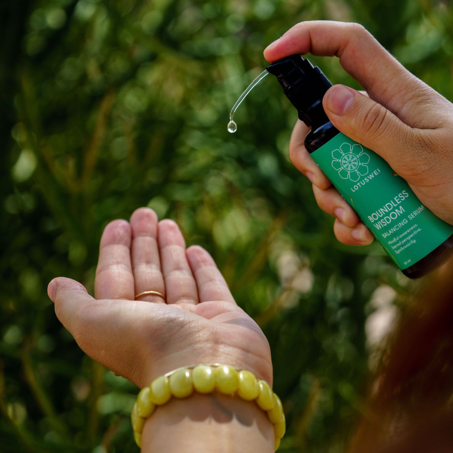 Hand receiving a drop of LOTUSWEI Boundless Wisdom serum from another hand, yellow bead bracelet visible, greenery behind