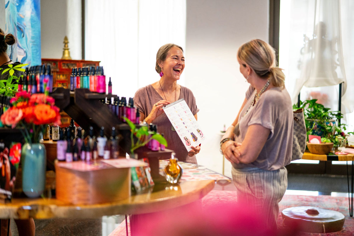 Two women laughing together in the LOTUSWEI shop, one holding a small color card, bottles and fresh flowers nearby